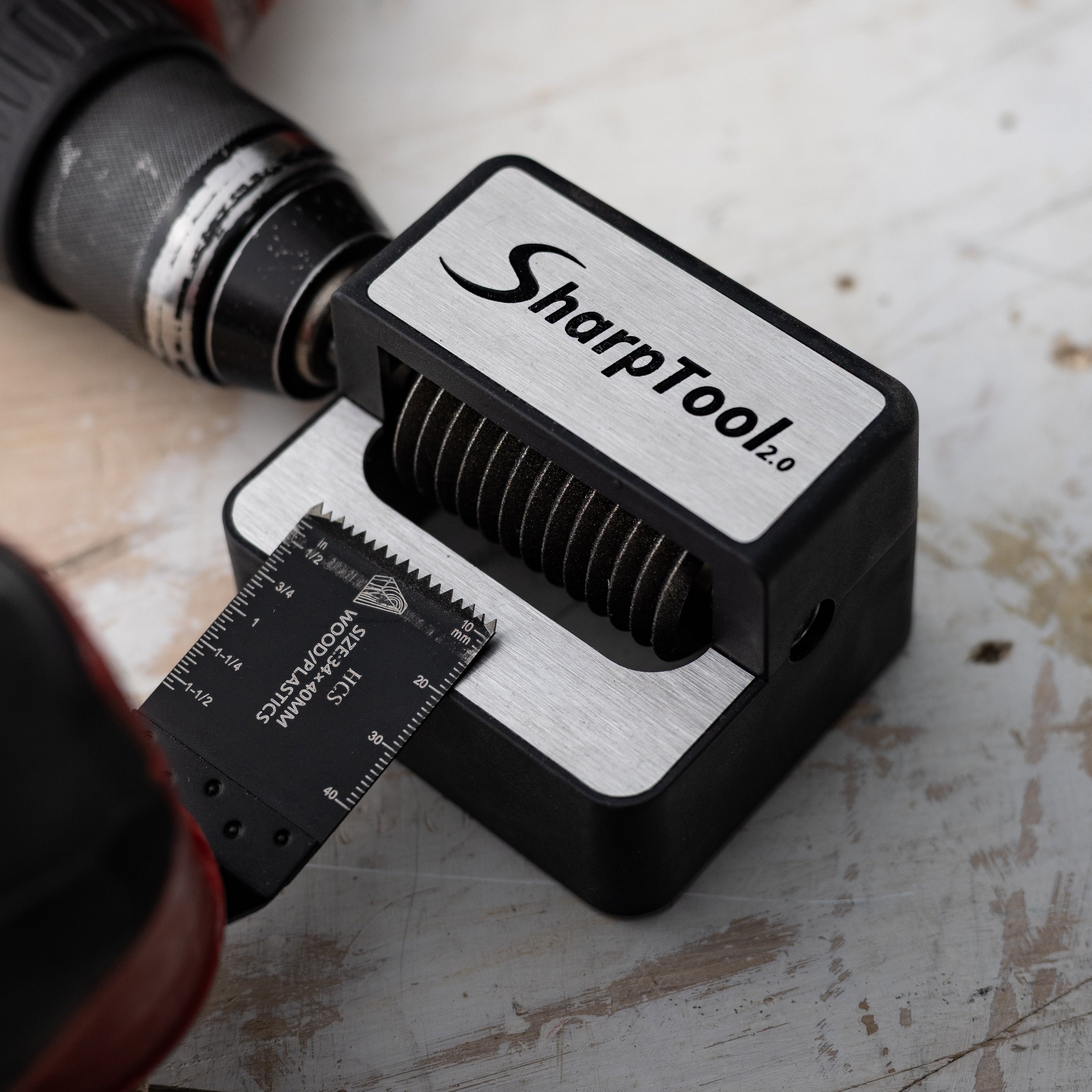 A SharpTool laser-etched steel square with a black and silver body, shown with a drill bit guide and part of a cordless drill on a worn, scratched white workbench surface.